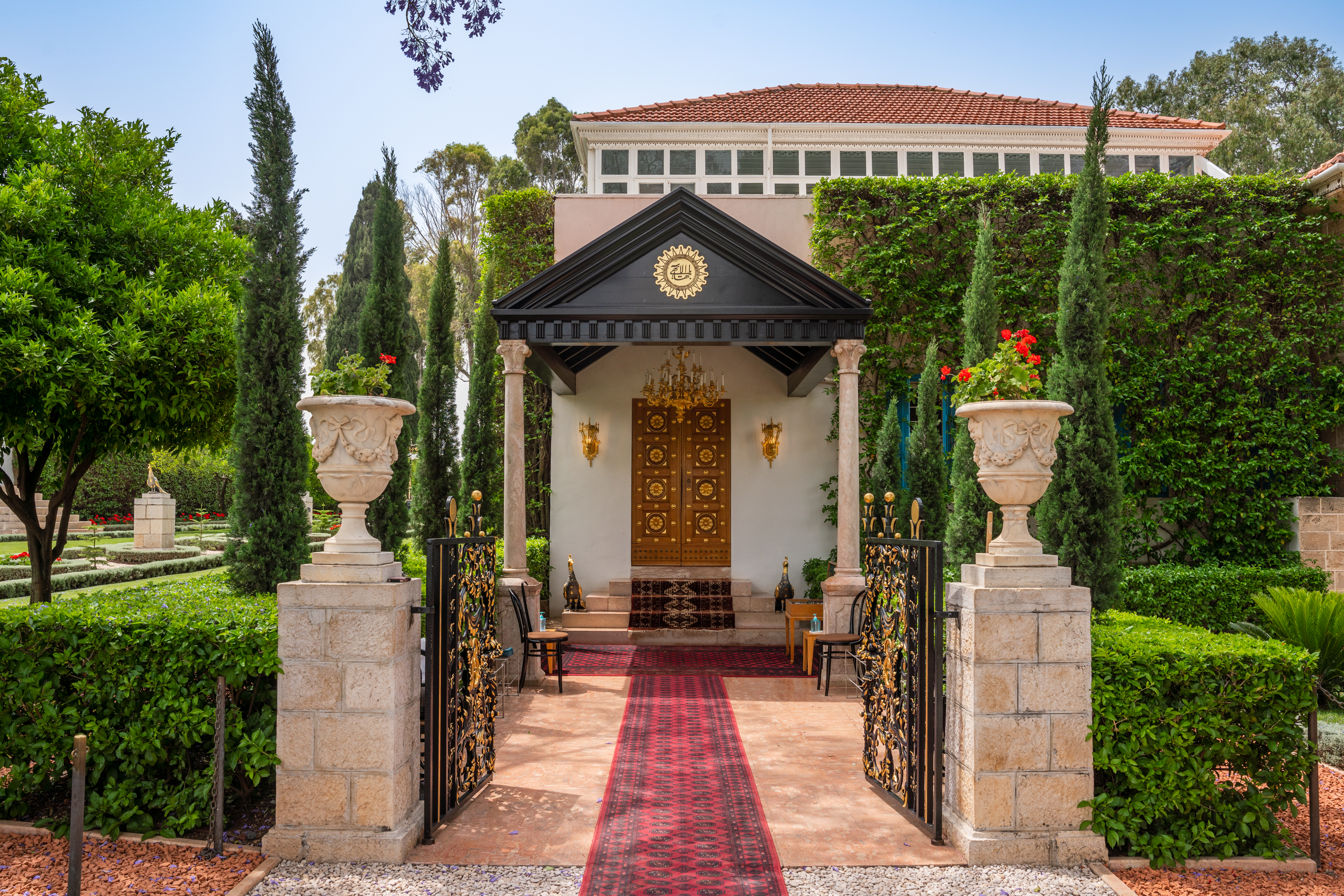 Las puertas doradas de un lugar sagrado Bahá'í, enmarcadas por cipreses y un sendero de jardín que conduce a la entrada.