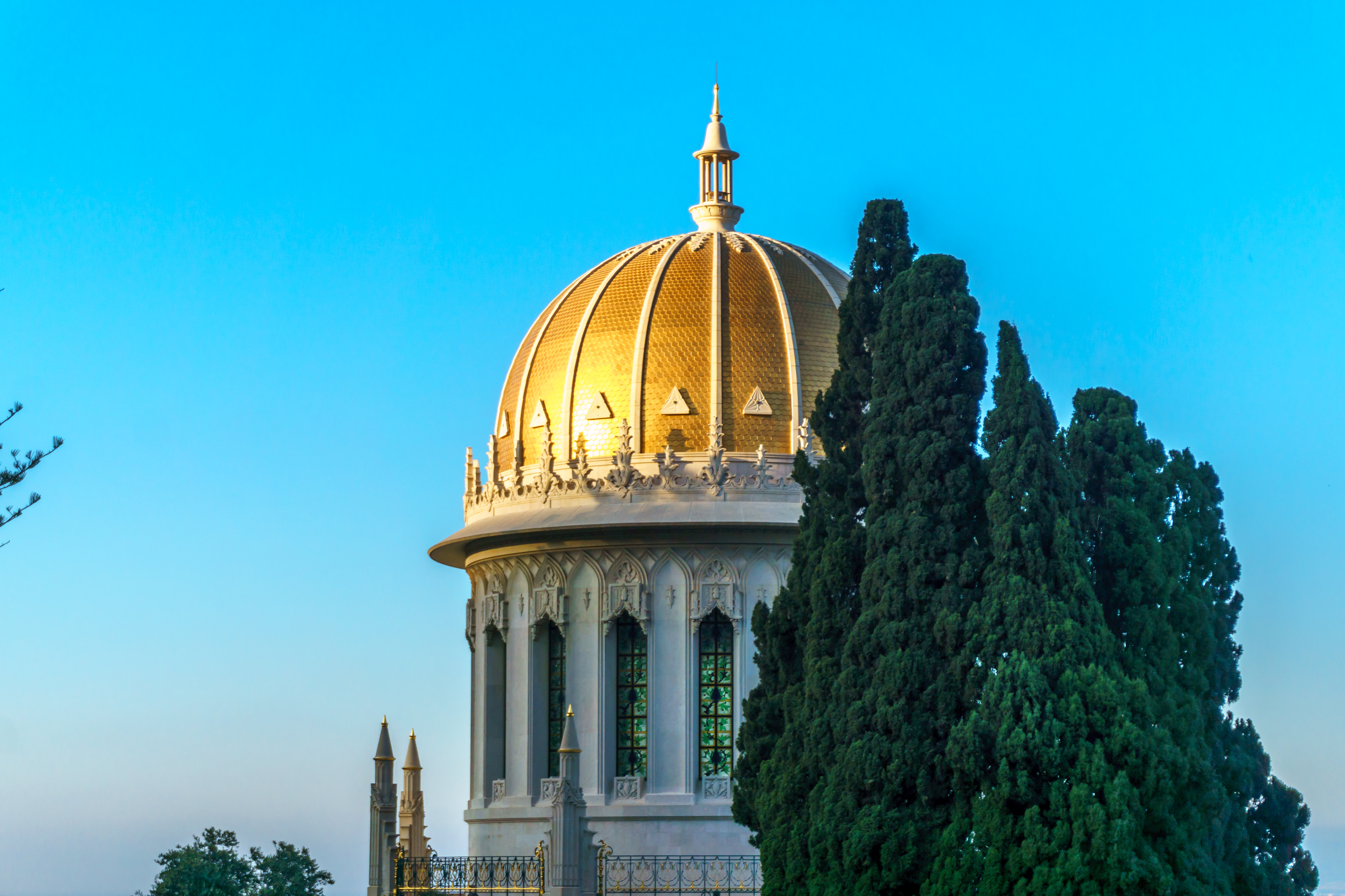 El Santuario del Báb — su cúpula dorada y sus vitrales elevándose contra un cielo azul, enmarcados por cipreses.